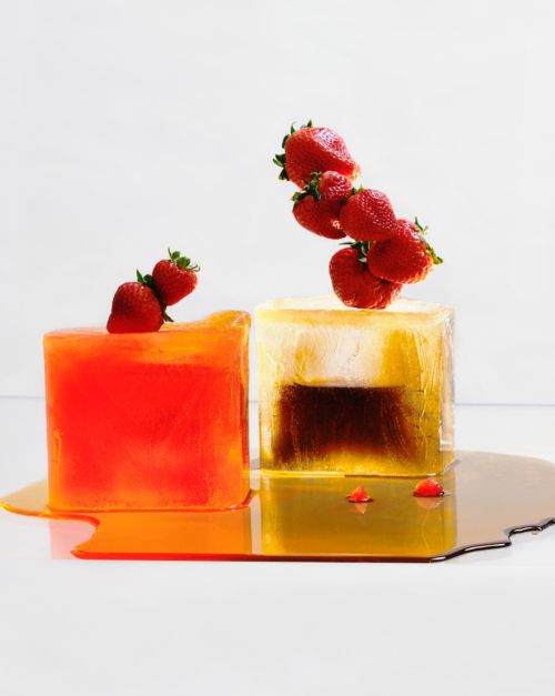 Two ice cubes topped with strawberries, one orange and one a gradient of brown to clear, melting on a white surface.