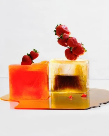 Two ice cubes topped with strawberries, one orange and one a gradient of brown to clear, melting on a white surface.