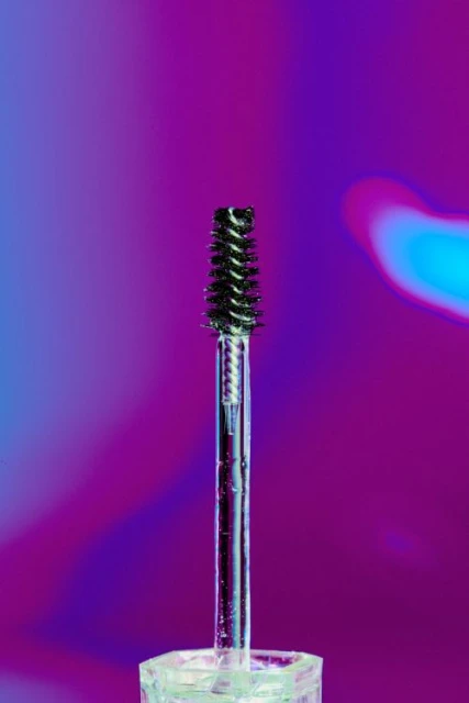 Close-up of a mascara wand with a twisted bristle brush against a purple and blue gradient background in a beauty context.