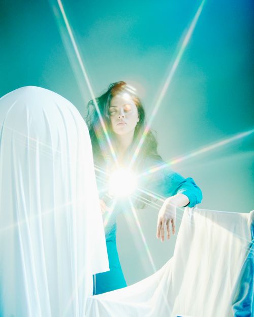 Woman in blue with eyes closed, surrounded by white fabric in a brightly lit indoor setting with strong starburst lens flares.