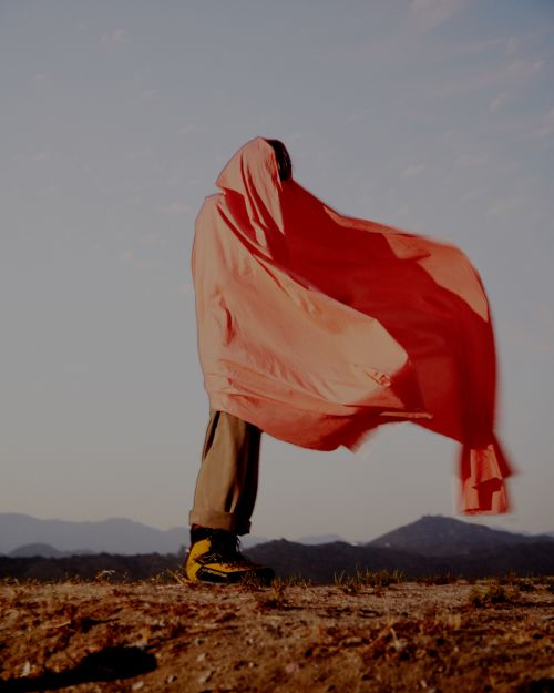 Person in a red hooded sheet standing outdoors. The figure wears tan pants and hiking boots with a landscape background under a light sky.