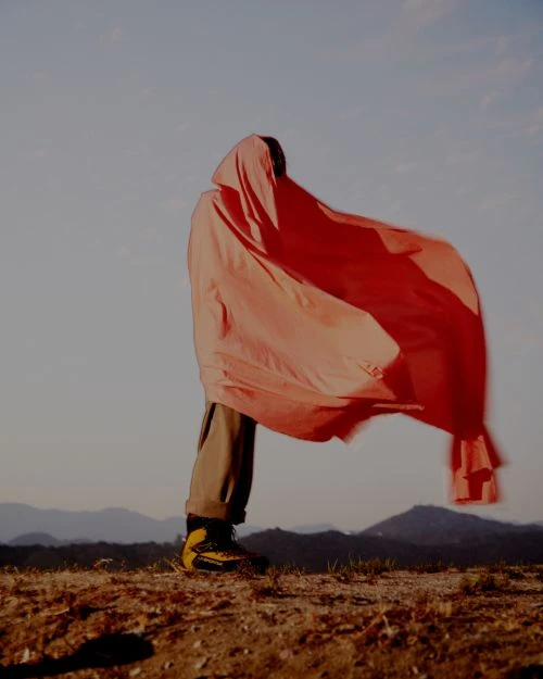 Person in a red hooded sheet standing outdoors. The figure wears tan pants and hiking boots with a landscape background under a light sky.