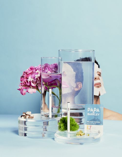 Still life with skincare face mask, clear glasses of water, Papa Barkley product and pink hydrangea flowers on a light blue table.