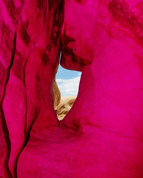 A vibrant close-up of a natural rock arch, featuring red rock formations and a glimpse of blue sky and tan rocks in the background.