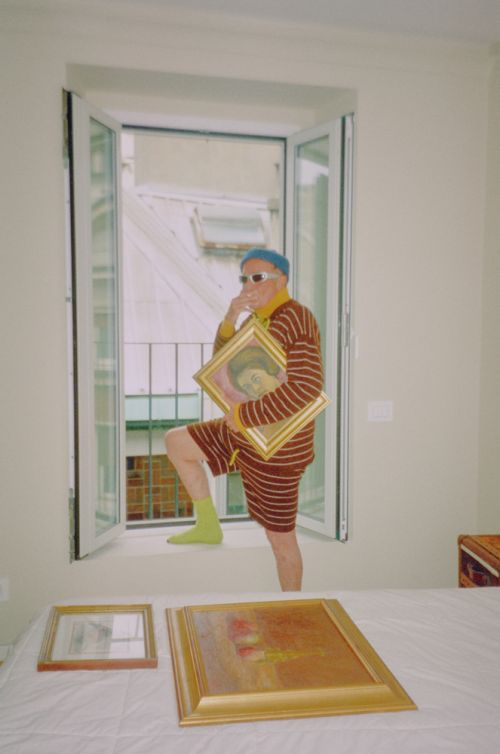 Elderly man in striped outfit poses in a window with art, standing above two framed paintings in a bright indoor setting.