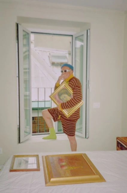 Elderly man in striped outfit poses in a window with art, standing above two framed paintings in a bright indoor setting.