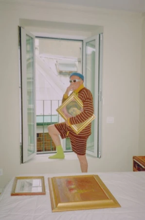 Elderly man in striped outfit poses in a window with art, standing above two framed paintings in a bright indoor setting.