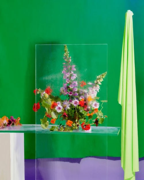 Still life with flowers in a vase on a glass shelf against a green background with a draped yellow cloth on the right.