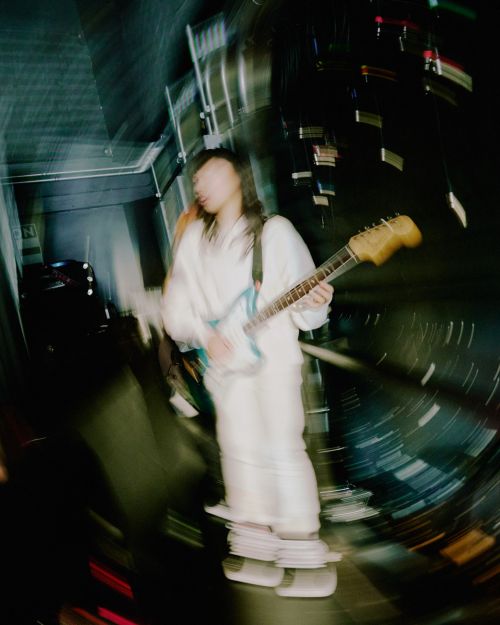 Unfocused shot of musician with long hair playing electric guitar in white outfit, indoor venue, stage lights, creative blur.