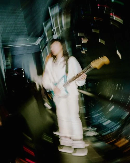 Unfocused shot of musician with long hair playing electric guitar in white outfit, indoor venue, stage lights, creative blur.
