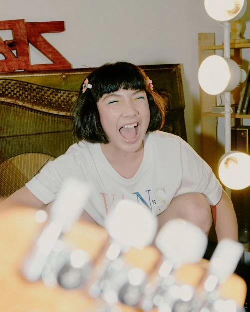 Girl with dark hair poses while grinning with a guitar in foreground and light panels in background, wearing a white T-shirt indoors.