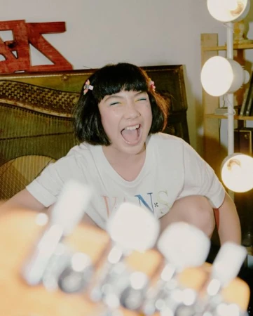 Girl with dark hair poses while grinning with a guitar in foreground and light panels in background, wearing a white T-shirt indoors.