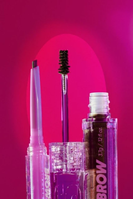 Close-up studio shot of brow products: pencil, mascara, and gel on a gradient pink background, highlighting the makeup's texture and color.