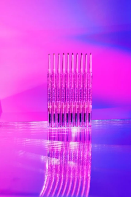 Stacked makeup brushes reflect vibrant neon light in a beauty still life against pink and blue backdrop, creating a bright, colorful abstract shot.