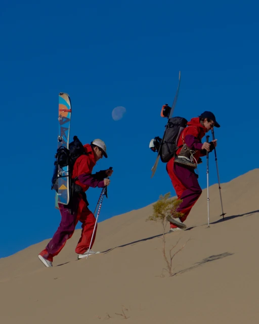 Two people hiking uphill in a desert landscape with skis and backpacks, under a clear blue sky with a pale moon visible.