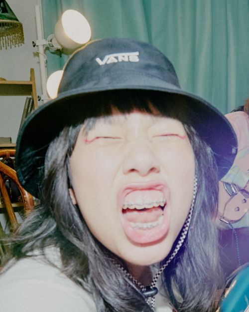 Close-up shot of a woman with open mouth and eyes closed, wearing a black Vans bucket hat with braces and a choker necklace indoors.