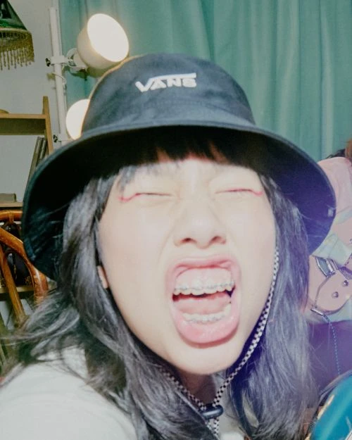 Close-up shot of a woman with open mouth and eyes closed, wearing a black Vans bucket hat with braces and a choker necklace indoors.