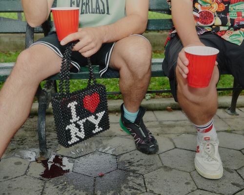Two men sit on a park bench holding red cups, with a beaded 'I love NY' purse on the ground, showcasing casual outdoor leisure.