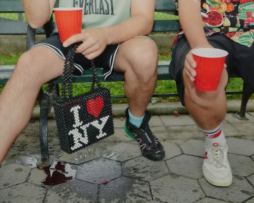 Two men sit on a park bench holding red cups, with a beaded 'I love NY' purse on the ground, showcasing casual outdoor leisure.