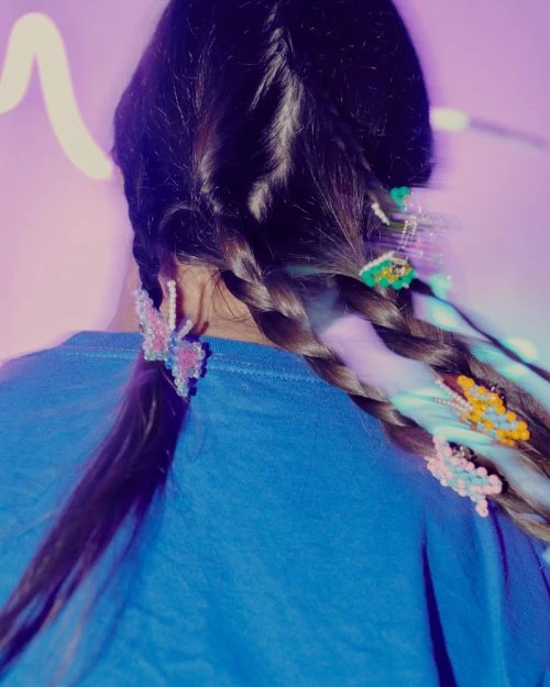 Rear view of a person with long brown hair styled in two braids adorned with colorful butterfly clips and a blue shirt on a light purple background.