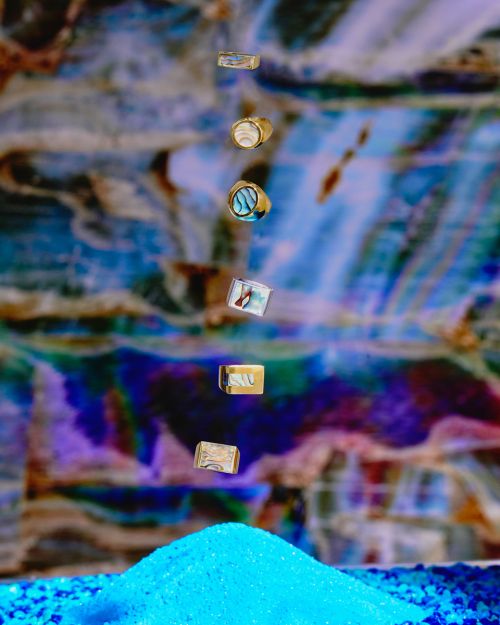 Falling gold rings cascade onto a blue sand pile against a blurred background featuring blue, white, purple, and brown hues.