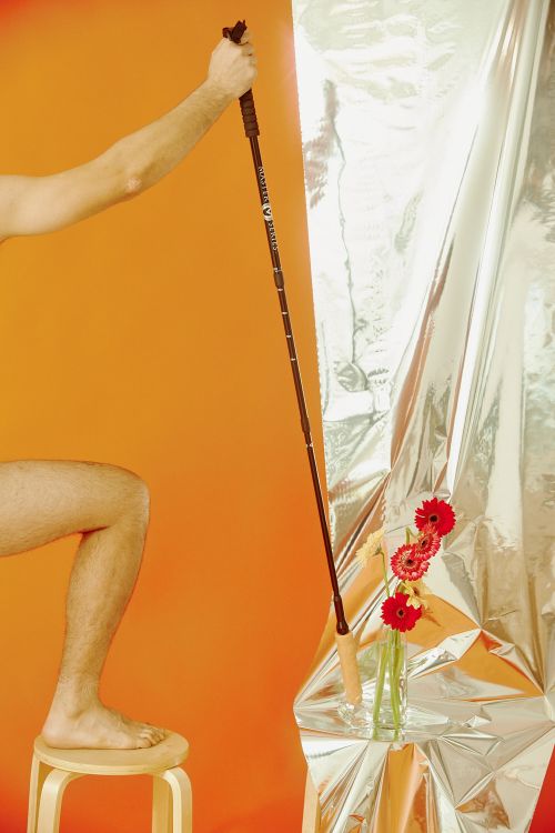 Person on stool holding trekking pole with red Gerbera daisies in vase against orange and silver background, studio lighting.