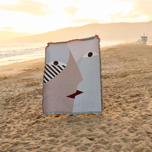 Abstract face art on a woven blanket on a sandy beach at sunset with a lifeguard tower in the distance and golden sky.