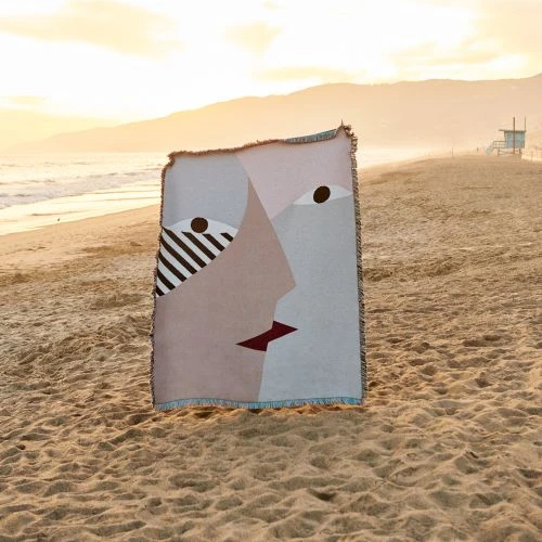Abstract face art on a woven blanket on a sandy beach at sunset with a lifeguard tower in the distance and golden sky.