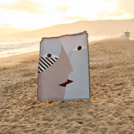 Abstract face art on a woven blanket on a sandy beach at sunset with a lifeguard tower in the distance and golden sky.
