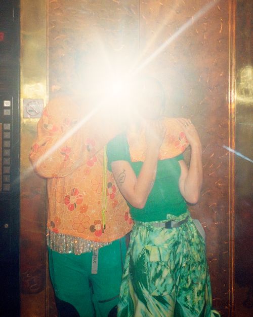 Two adults pose indoors against a gold-toned wall. One is in an orange floral top and green pants, and the other is in green separates.