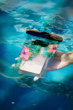Hand holds a waterproof phone pouch with floral designs, suspended in blue water with light reflections, demonstrating water protection.