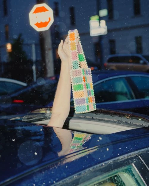 A woman's arm extends out of a car sunroof holding a beaded bag in a nighttime city scene with a stop sign in the background.