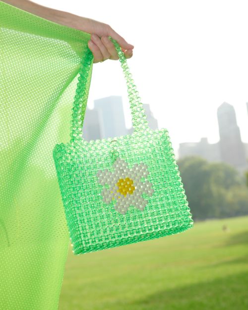 Close-up of a green beaded handbag with a daisy design, held by a hand, against a backdrop of blurred city buildings and green grass.