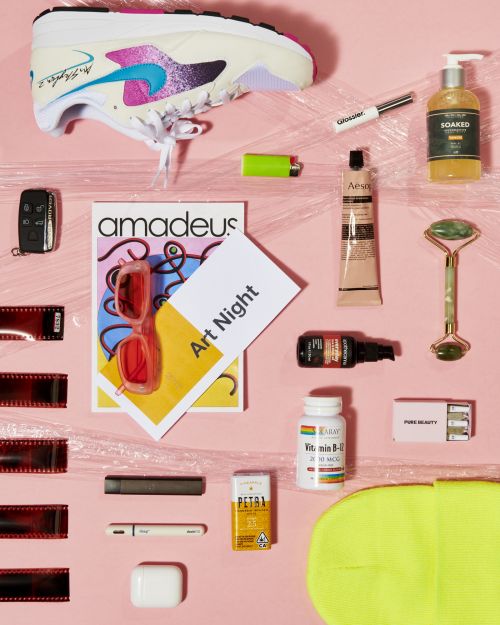 Flat lay of a sneaker, skincare products, film rolls, and accessories arranged on a pink background covered in clear plastic wrap.