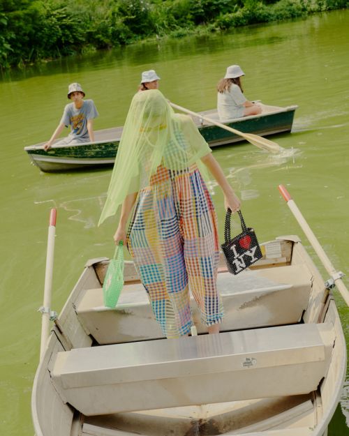 Four people in rowboats on a pond with trees, wearing casual clothing and hats, carrying shopping bags, and enjoying a sunny day outdoors.