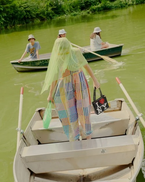 Four people in rowboats on a pond with trees, wearing casual clothing and hats, carrying shopping bags, and enjoying a sunny day outdoors.