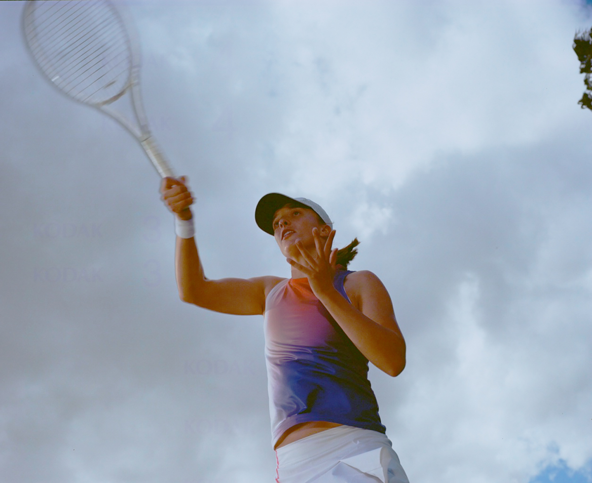 Female tennis player with a racquet raised against a cloudy sky, wearing a blue and pink tank top and visor. Focus on sports and athleticism.