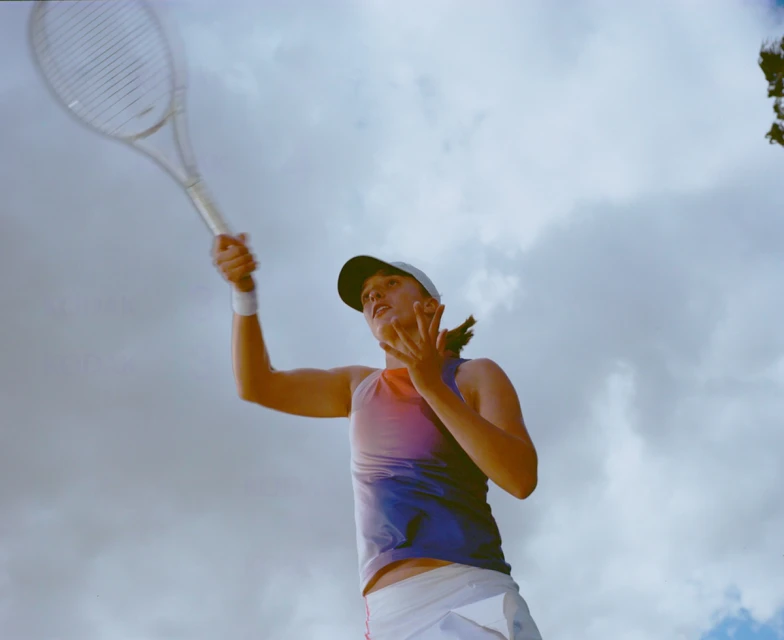Female tennis player with a racquet raised against a cloudy sky, wearing a blue and pink tank top and visor. Focus on sports and athleticism.