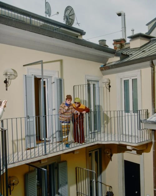 Two people on a balcony of a yellow building wear striped tops and hats, with one leaning on railing and the other smoking.