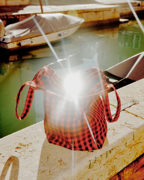 Red and black checkered patterned bucket bag with drawstring closure sitting on a stone ledge in a canal setting with boats visible.