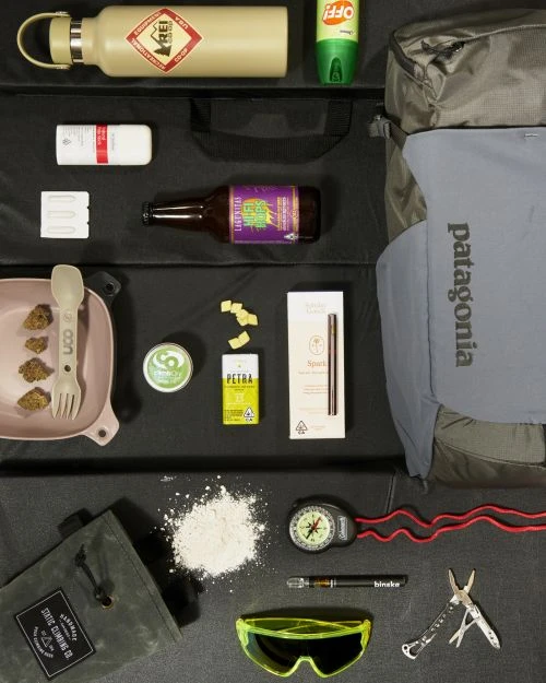 Flat lay of outdoor gear including a water bottle, bug spray, backpack, snacks, compass, and safety glasses on a black background.