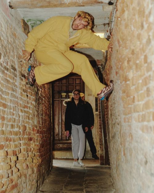 Adult male in yellow suit climbing brick alleyway with two people standing by a door in the background looking up in an urban setting.