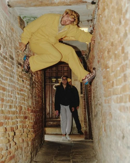 Adult male in yellow suit climbing brick alleyway with two people standing by a door in the background looking up in an urban setting.