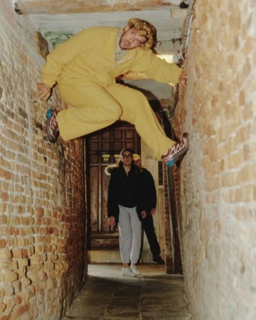 Adult male in yellow suit climbing brick alleyway with two people standing by a door in the background looking up in an urban setting.