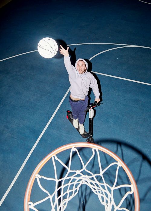 Eye-level shot of a man on a scooter, aiming a basketball toward a hoop with a net on an outdoor blue basketball court at night.