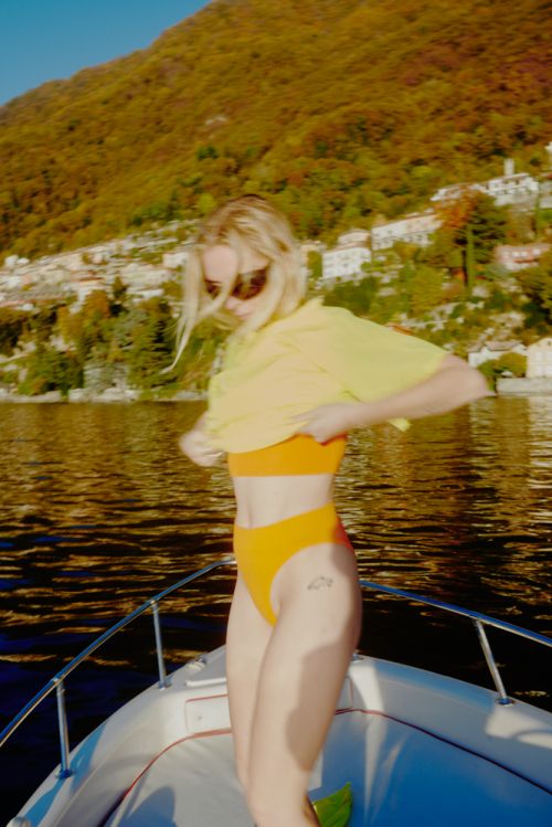 Woman on a boat removing yellow shirt to reveal orange bikini top and bottoms against a backdrop of a lake and hillside.