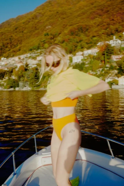 Woman on a boat removing yellow shirt to reveal orange bikini top and bottoms against a backdrop of a lake and hillside.
