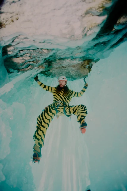 A man ice climbing on a frozen waterfall in a yellow and black striped suit, wearing a helmet and climbing gear on a sunny day.