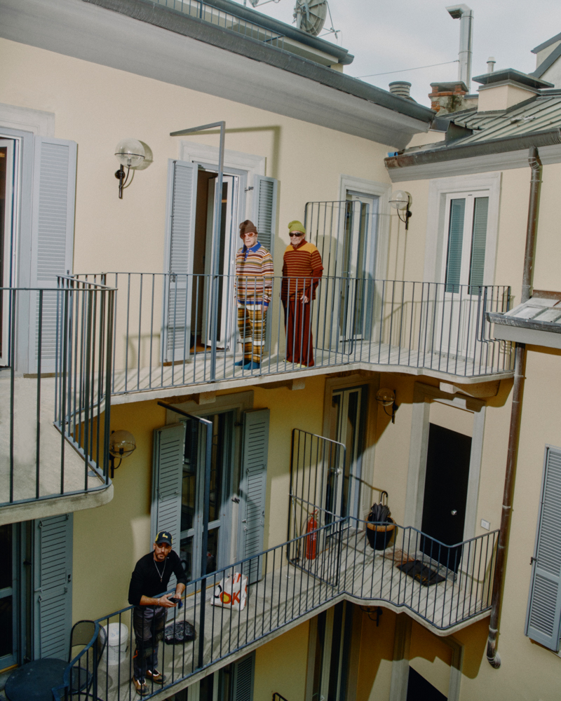 Three-story building with balconies features three people in colorful outfits on the top balconies, and one person on the lower balcony.