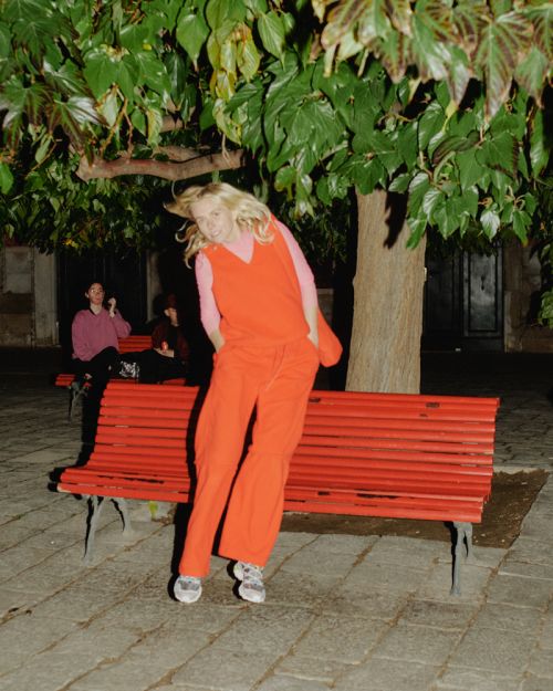 Woman in orange suit standing in front of a red bench with green trees in the background and two people sitting on the bench.